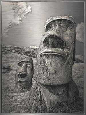 An intricate stainless steel etching captures two moai statues on a windswept hillside, rendered in silver-gray crosshatching and dark recessed shadows. The foreground figure dominates with weathered texture and solemn expression, while a smaller statue rests behind. Brushed metal sky, clouds, grass, and beveled border give refined depth and elegance.