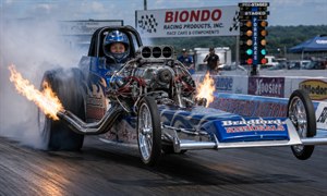 An experienced female racer launches a blue-and-silver dragster just beyond the starting line. Wearing a matching helmet, she grips the cockpit as the exposed supercharged engine roars. Flaming exhaust pipes, spinning rear tires, heavy smoke, blurred pavement, sponsor barriers, and staging lights create a dramatic, high-speed motorsport moment of acceleration.