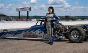 A confident female drag racer stands beside a sleek blue-and-silver dragster at a sunny speedway. Wearing a detailed racing suit and holding a matching helmet, she smiles near the exposed chrome engine. Sponsor signs, grandstands, timing boards, enormous slick tires, and bright clouds complete the powerful motorsport portrait with realism.