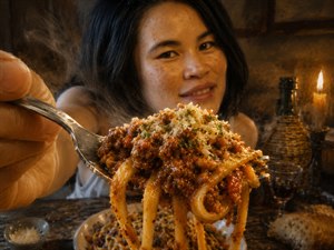 A warm candlelit moment captures a smiling woman offering an abundant forkful of glossy pasta and rich meat sauce. Parmesan, herbs, and rising steam heighten the delicious realism, while the weathered wooden table, rustic bread, wine, and soft background create an intimate, inviting dining atmosphere filled with comfort and generosity.