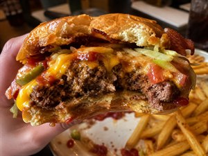 A towering cheeseburger bursts with juicy beef, melted cheese, crisp bacon, lettuce, tomato, pickles, onions, and glossy sauces spilling from every side. Held close to the viewer after several bites, it reveals rich textures and dripping flavor, while golden fries and a messy plate complete the indulgent, realistic dinner scene.