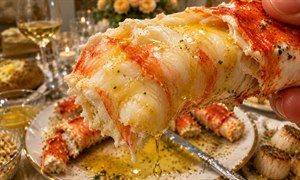An extreme close-up captures a tender piece of Alaskan king crab lifted toward the diner, its shell mostly removed to reveal delicate, richly textured meat. Golden butter glistens across every strand and drips toward the elegant plate below, surrounded by crab legs, scallops, baked potato, and warm candlelit dining details.