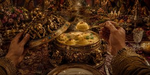 An opulent fantasy banquet unfolds beneath moonlit temple towers, where a jeweled ceremonial vessel releases curling steam. First-person hands lift its gilded lid and raise a spoonful of mysterious delicacy. Candlelight gleams across carved monkey motifs, silverware, crystal, fruit, embroidered cloth, and lavish dishes, creating vintage adventure intrigue and suspense.