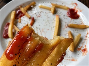 Crispy golden French fries fill the frame in a mouthwatering extreme closeup, glistening with rich ketchup, coarse salt, and cracked black pepper. A freshly bitten fry reveals its fluffy center, while the nearly finished plate behind adds irresistible realism. Bold, savory, satisfying artwork created to awaken cravings instantly and appetites.