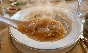 An elegant dining scene captures a steaming spoonful of shark fin soup approaching the viewer. Glossy amber broth clings to delicate translucent strands, rendered in mouthwatering detail. Behind it, the ornate bowl sits mostly empty, surrounded by refined tableware, soft lighting, warm reflections, and a truly luxurious restaurant atmosphere throughout.