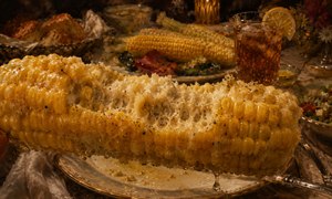 Golden butter glistens across a mostly eaten corn cob, held close from an immersive diners viewpoint. Detailed kernels, rough bite marks, pepper, salt, and dripping butter dominate the foreground, while elegant china, iced tea, roasted vegetables, bread, and softly lit table settings create a warm, luxurious painted feast behind it.