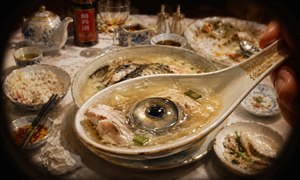 An extreme close-up captures a porcelain spoon approaching the viewer, brimming with rich fish-head broth, tender flakes, herbs, and a striking glossy eye. Beyond it, the half-eaten soup bowl and elegant banquet table fade softly into view, creating an immersive, dramatic, richly painted dining perspective filled with texture and atmosphere.