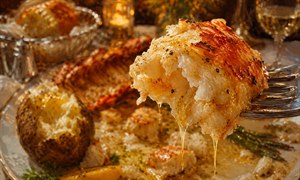 An irresistible forkful of tender lobster fills the foreground, glistening beneath warm restaurant light as golden butter streams from every succulent fibre. Behind it, a half-finished seafood feast of baked potato, scallops, asparagus, lemon, and lobster shell softens into a luxurious candlelit blur, capturing pure fine-dining indulgence and anticipation beautifully.