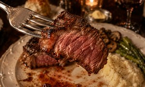Succulent medium-rare steak fills the foreground, lifted toward the viewer on a polished fork, its seared crust glistening with rich juices. Behind it, a half-finished plate of creamy potatoes, asparagus, mushrooms, and steak rests beneath warm candlelight, creating an intimate, mouthwatering celebration of fine dining and irresistible flavor in vivid detail.