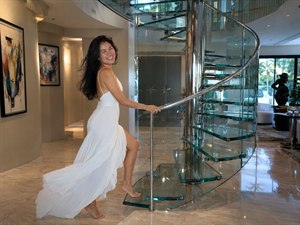 A graceful woman in a flowing white gown ascends a sculptural glass spiral staircase inside a luminous modern mansion. Her windswept black hair and billowing skirt add romantic movement, while mirrored chrome, polished marble, warm lighting, abstract art, and garden views create an elegant cinematic atmosphere of luxury and serenity.