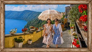 Two lifelong friends stroll arm in arm along a sunlit Italian lakeside path, sharing an elegant floral parasol beneath a brilliant summer sky. Rustic stonework, vivid geraniums, blue water, forested hills and hillside homes surround them, creating a warm, joyful portrait of friendship, travel and timeless Mediterranean beauty together forever.