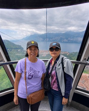Two lifelong friends pose inside a panoramic Italian cable car, framed by sweeping windows and rugged mountain scenery. Below, a red-roofed lakeside village rests beside calm blue water. Their relaxed smiles, travel clothes, and close companionship capture a joyful sightseeing moment high above the beautiful landscape of northern Italy together.