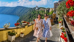 Two lifelong friends stroll arm in arm along a sunlit Italian hillside path, surrounded by brilliant geraniums, rustic stonework, and breathtaking lake views. Elegant Mediterranean dresses, warm smiles, and relaxed companionship capture the joy of travel, friendship, and unforgettable moments shared beneath Italys radiant summer sky in timeless beauty forever.