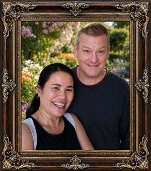 A cheerful couple poses closely together in a vibrant botanical garden, surrounded by softly blurred blossoms and lush greenery. Their warm smiles create an affectionate, relaxed mood. An ornate dark oak frame, enriched with gold and chrome inlays and elaborate sculpted corners, gives the portrait an elegant heirloom finish appearance.