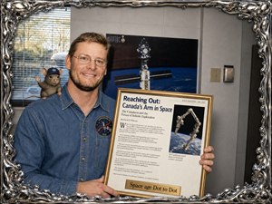 A warm, confident portrait shows a smiling man in a detailed denim shirt holding a gold-framed magazine-style award titled Space age Dot to Dot. The article celebrates Canadas arm in space, with a Canadarm image and simulated text. A tiny mouse wearing a NASA-style cap waves from his shoulder, all surrounded by a shiny mercury-wax frame.