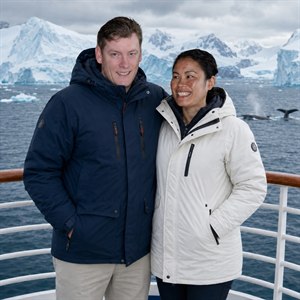 An adventurous couple stands warmly together on a polar ship deck, wrapped in rugged parkas beside polished railings. Behind them, icy waters, towering blue-white icebergs, stormy mountains, and distant whales create a breathtaking arctic voyage. Crisp faces, bright smiles, and cinematic detail make this portrait feel romantic, bold, and unforgettable.