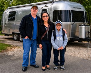 A happy family stands together in front of a polished aluminum travel trailer, ready for adventure. The bright outdoor scene features crisp details, warm smiles, reflective metal, casual clothing, and a natural wooded setting. It feels cheerful, memorable, and perfect for celebrating travel, family bonding, and lasting vacation memories.