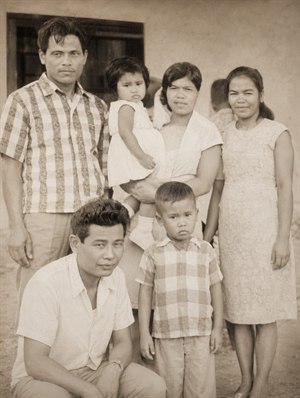 Restored sepia family portrait showing six people posed outdoors in front of a simple building wall and dark doorway. The photo has repaired cracks, softened stains, improved contrast, and clearer faces. A man stands left, a woman stands right, a central woman holds a toddler, with a young boy and crouching man in front.