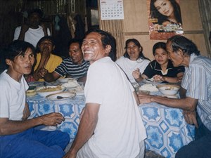 Restored with clearer detail and warmer balanced light, this candid family meal captures laughter, conversation, and togetherness around a busy table. Bowls of rice, patterned cloth, bamboo walls, calendar, and poster frame the scene, while sharpened faces, natural colors, and softened haze preserve the heartfelt charm of a treasured memory.