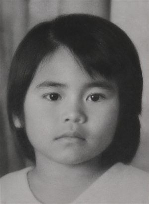 A restored black-and-white close-up portrait of a young child facing forward with a calm, serious expression. The image has soft vintage lighting, smooth grayscale tones, dark straight hair framing the face, clear eyes, and a simple blurred background. The repaired photo preserves an old studio-style, nostalgic feeling.