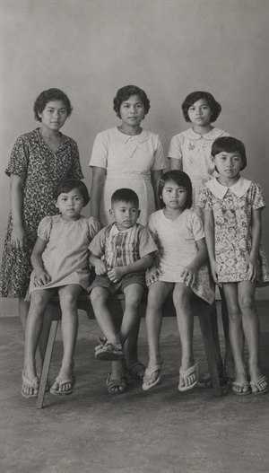 Restored vintage black-and-white family portrait showing seven people posed formally in a simple studio setting. The cleaned image reveals clearer faces, clothing textures, sandals, bench details, and soft floor shadows. The wider framing gives the portrait more breathing room while preserving the quiet, nostalgic character of the original photograph.