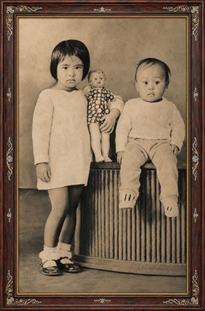 A beautifully restored sepia portrait captures two young children in a timeless studio pose. The rosewood frame, with gold inlay and ornate completed corners, adds antique elegance. Soft tones, crisp details, knitted clothing, flowers, and calm expressions create a cherished family keepsake full of warmth, history, and gentle nostalgic beauty.